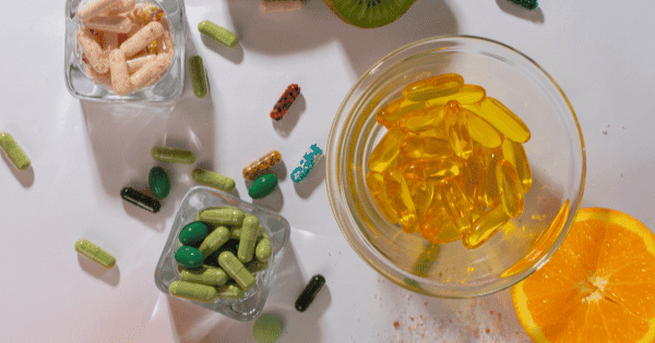 A variety of colorful supplement capsules, softgels in a glass bowl, and sliced citrus fruit on a white background.