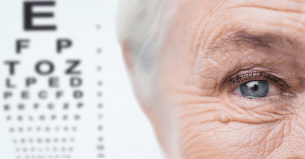 A close-up of an elderly person's blue eye on the right side of the image, with a blurred Snellen eye test chart visible in the background on the left.