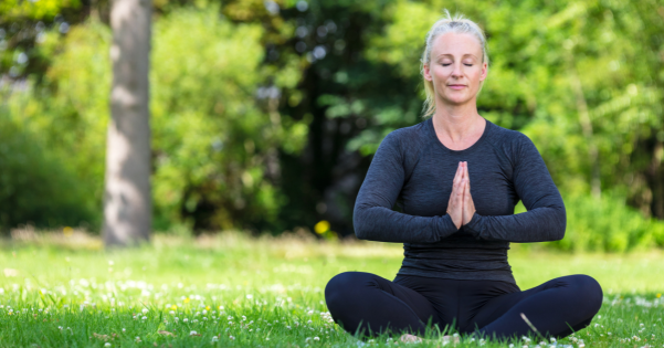 A woman sitting in a cross-legged yoga position outdoors on grass with her hands together in prayer.
